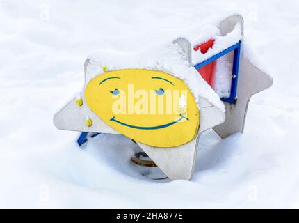 Snowy playground in winter, Moscow, Russia. Smile image on children`s swing, empty park after snowfall. Scenery of kindergarten under snow in winter c Stock Photo